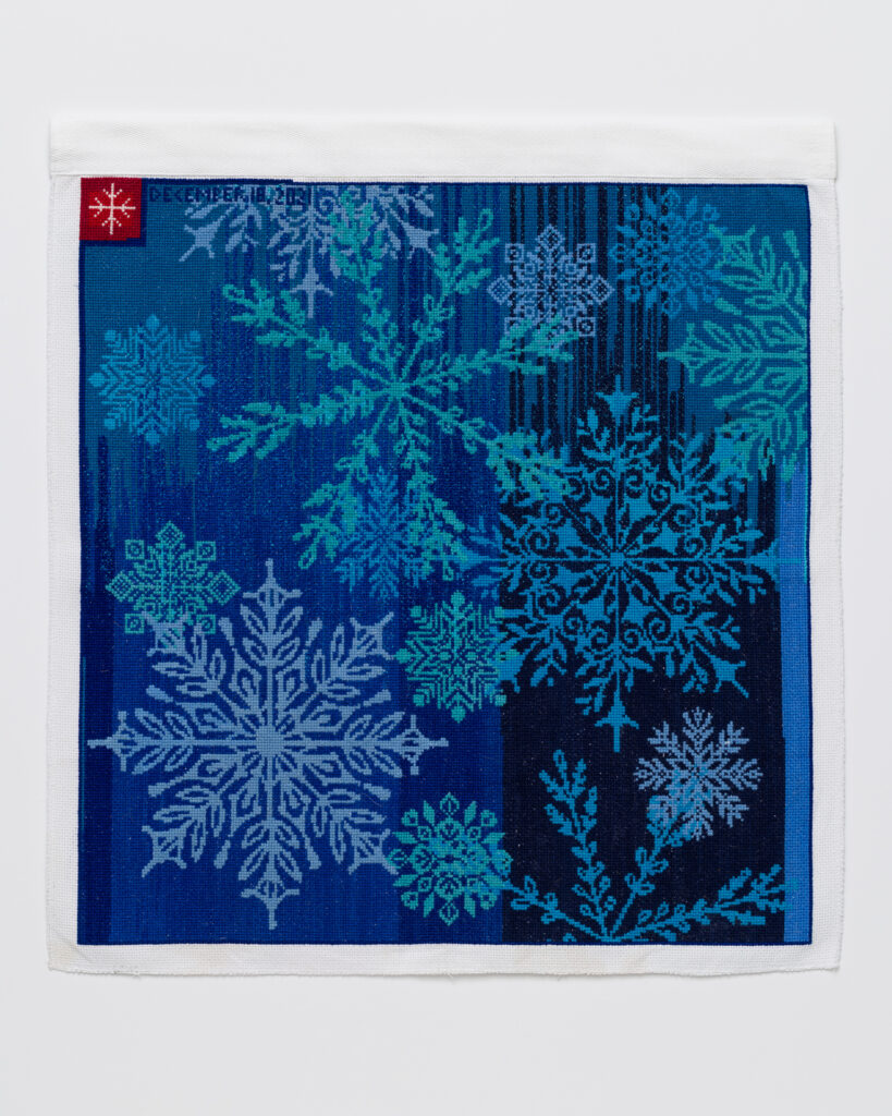 Cross stitch embroidery with turquoise and light blue snow flakes on a medium and dark blue background. A small red square is in the top left corner with a small pink snowflake inside.