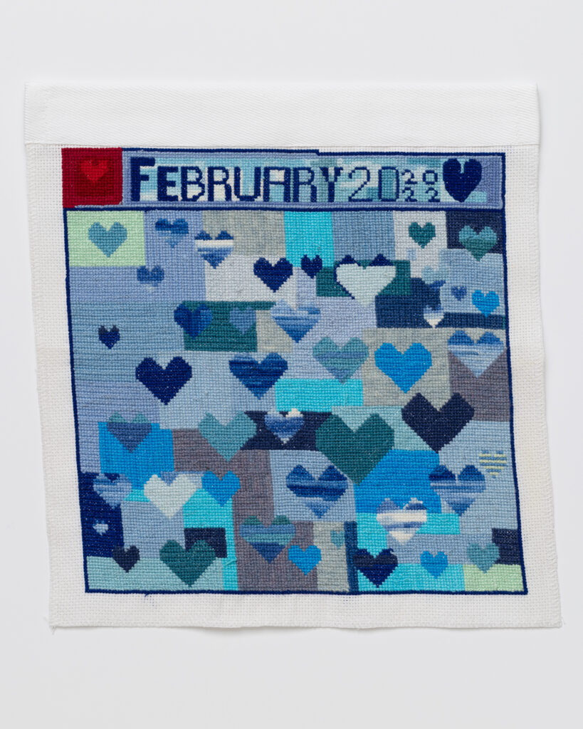 Cross stitch embroidery with various colored blue hearts on a background of vlue rectangles. 