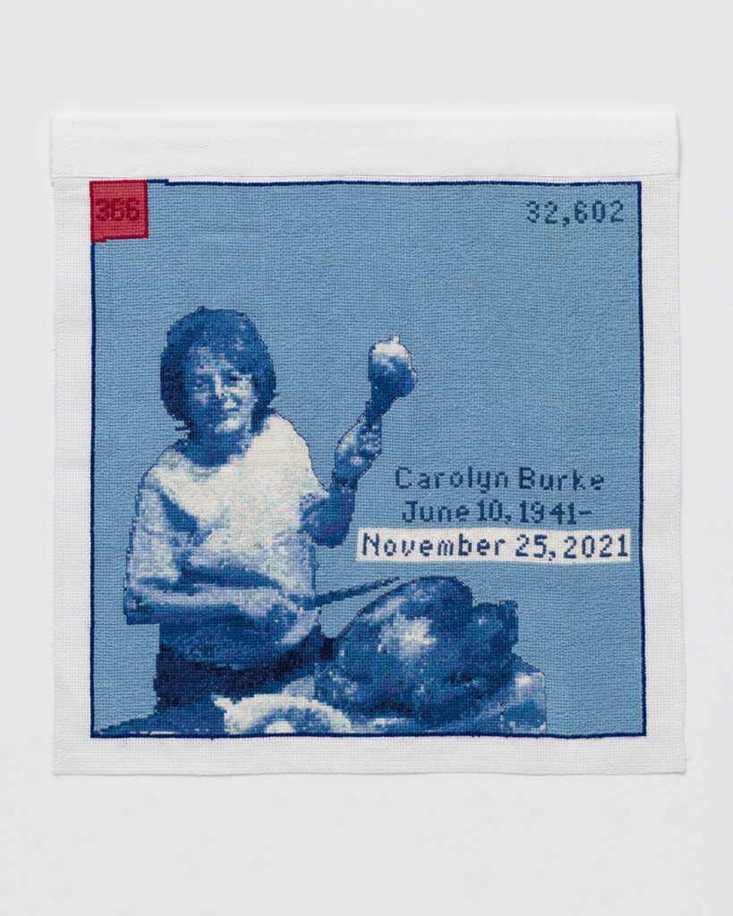 Cross stitch embroidery of a woman with short hair in a plain t-shirt holding up a turkey leg in her left hand, with a knife in her right hand. A cooked turkey sits on a table in front of her. Stitched in blue threads, with a red square in the top left corner. Across the middle right of the image is written 