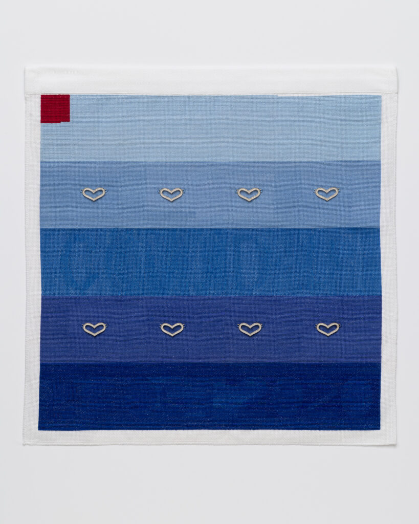 Cross stitch embroidery of 5 sections of blue colors from light to dark, top to bottom. 8 silver hearts are attached across the 2nd and 4th sections. There is a red square in the top left corner.