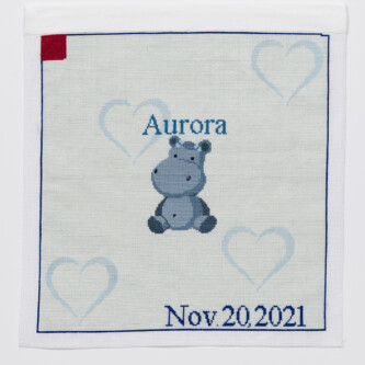 Photo by Wes Magyar Cross stitch embroidery of a cartoon hippopotamus with the name "Aurora" stitched above it. The background is very light blue with light blue hearts. "Nov 20, 2021" is stitched on the bottom right corner, and a red square is in the top left.