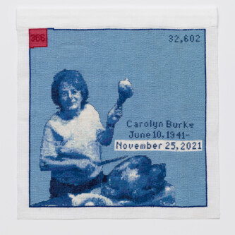 Photo by Wes Magyar Cross stitch embroidery of a woman with short hair in a plain t-shirt holding up a turkey leg in her left hand, with a knife in her right hand. A cooked turkey sits on a table in front of her. Stitched in blue threads, with a red square in the top left corner. Across the middle right of the image is written "Carol Burke June 10, 1941 - November 25, 2021"