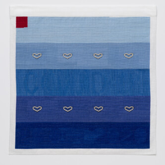 Cross stitch embroidery of 5 sections of blue colors from light to dark, top to bottom. 8 silver hearts are attached across the 2nd and 4th sections. There is a red square in the top left corner.