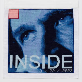 Photo by Wes Magyar Cross stitch embroidery of the top half of a man's face in various colors of blue, with the eyes looking left. There is a small pink square outlined in red thread in the top left corner. Across the bottom of the image is the word "INSIDE" in light blue thread.