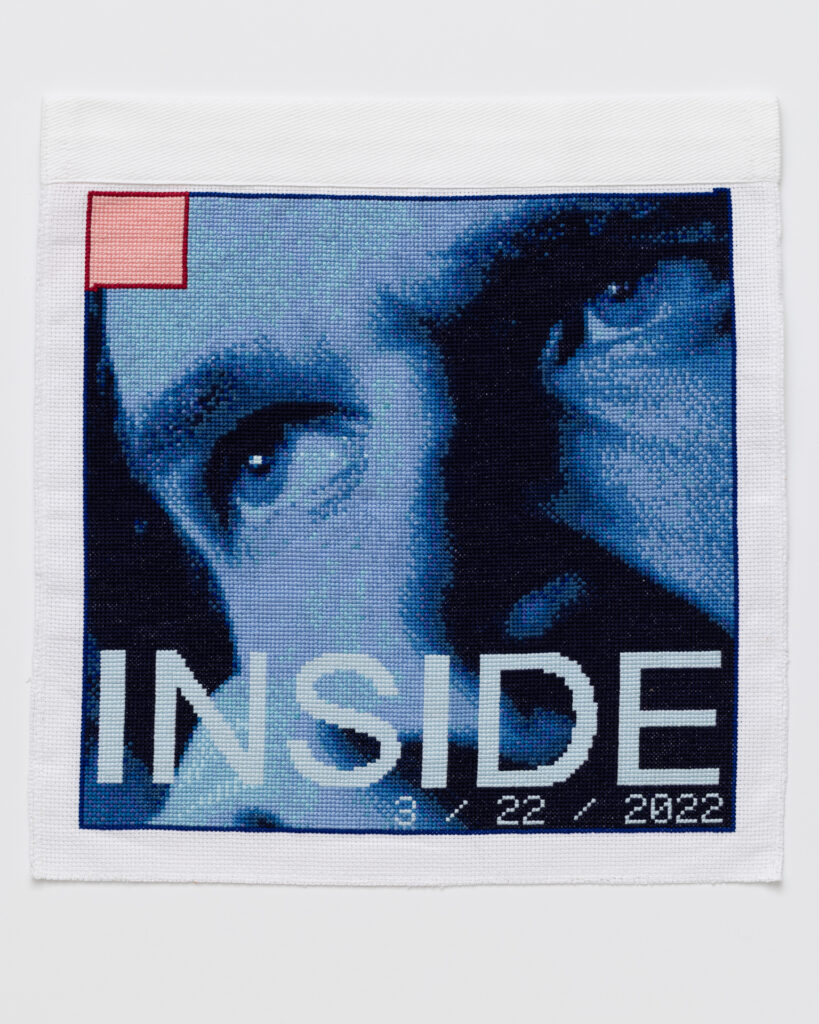 Cross stitch embroidery of the top half of a man's face in various colors of blue, with the eyes looking left. There is a small pink square outlined in red thread in the top left corner. Across the bottom of the image is the word 